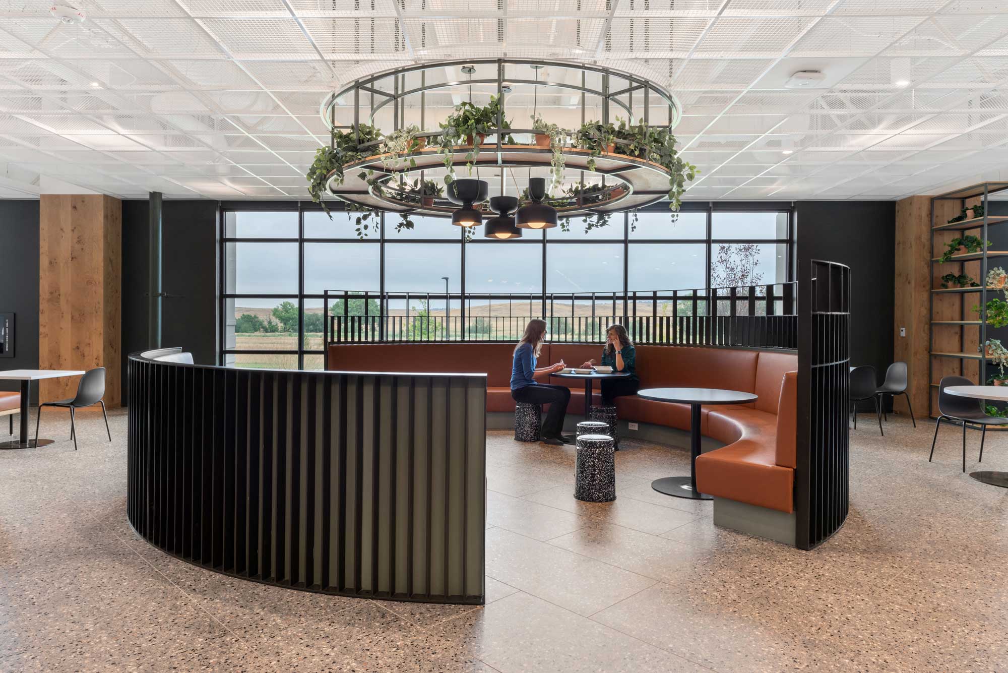Two employees meet in a curved leather booth at the Medtronic Lafayette Campus, beneath a circular chandelier draped with hanging plants, with floor-to-ceiling…Two employees meet in a curved leather booth at the Medtronic Lafayette Campus, beneath a circular chandelier draped with hanging plants, with floor-to-ceiling windows overlooking open Colorado plains.
