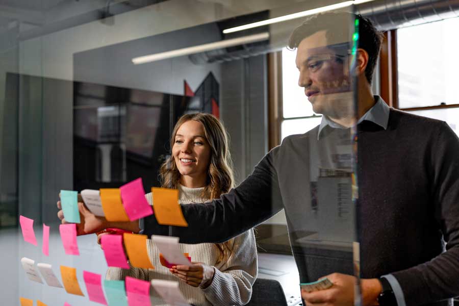 > `Two MOA Architecture team members organizing colorful sticky notes on a glass wall during a collaborative brainstorming session.