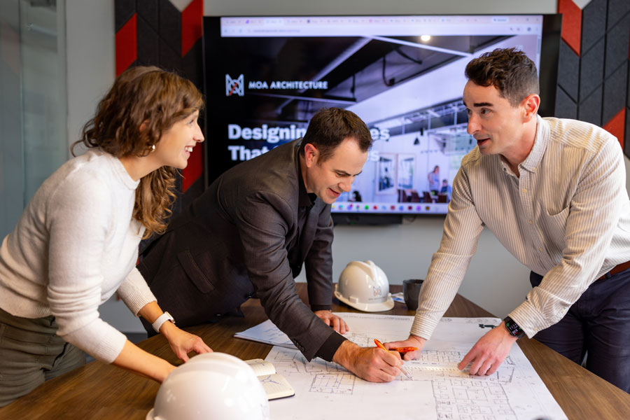 > `Three MOA Architecture team members collaborating over blueprints at a conference table with an MOA presentation visible on screen behind them.