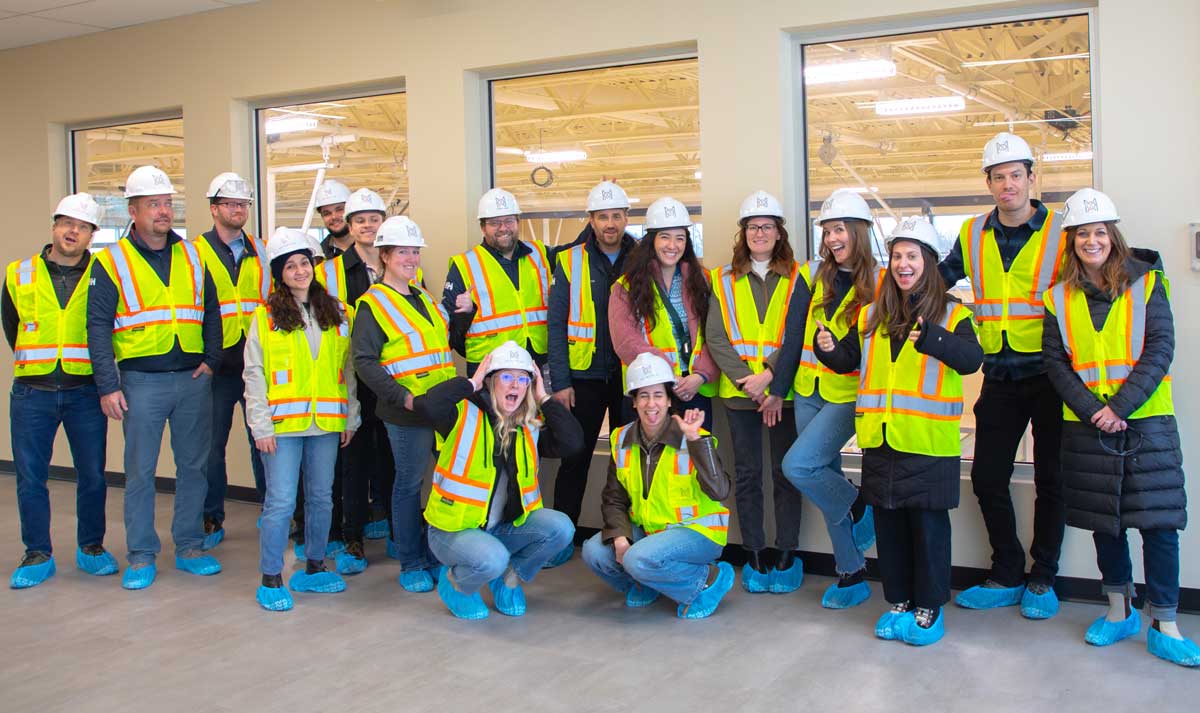 The MOA Architecture team smiling together on a construction site walkthrough, wearing hard hats, yellow safety vests, and blue protective shoe covers.