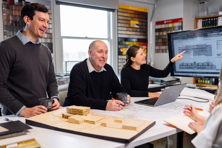> `MOA Architecture team members gathered around a studio table with a physical scale model and blueprints, reviewing architectural drawings on a large monitor.