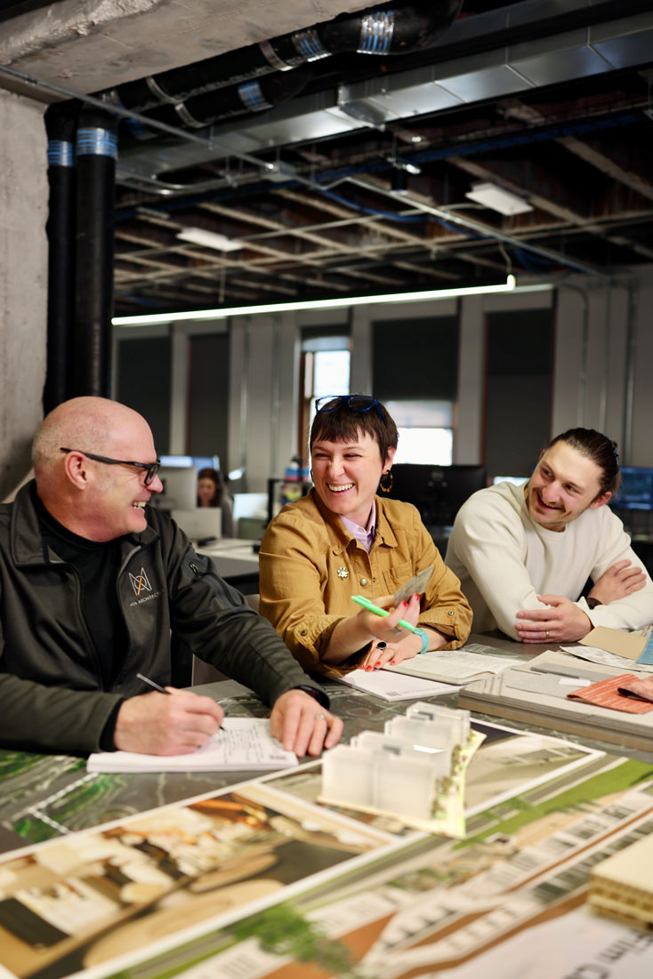 Three MOA Architecture team members laughing together during a design review, with project renderings, a small building model, and notes spread across the studio table.
