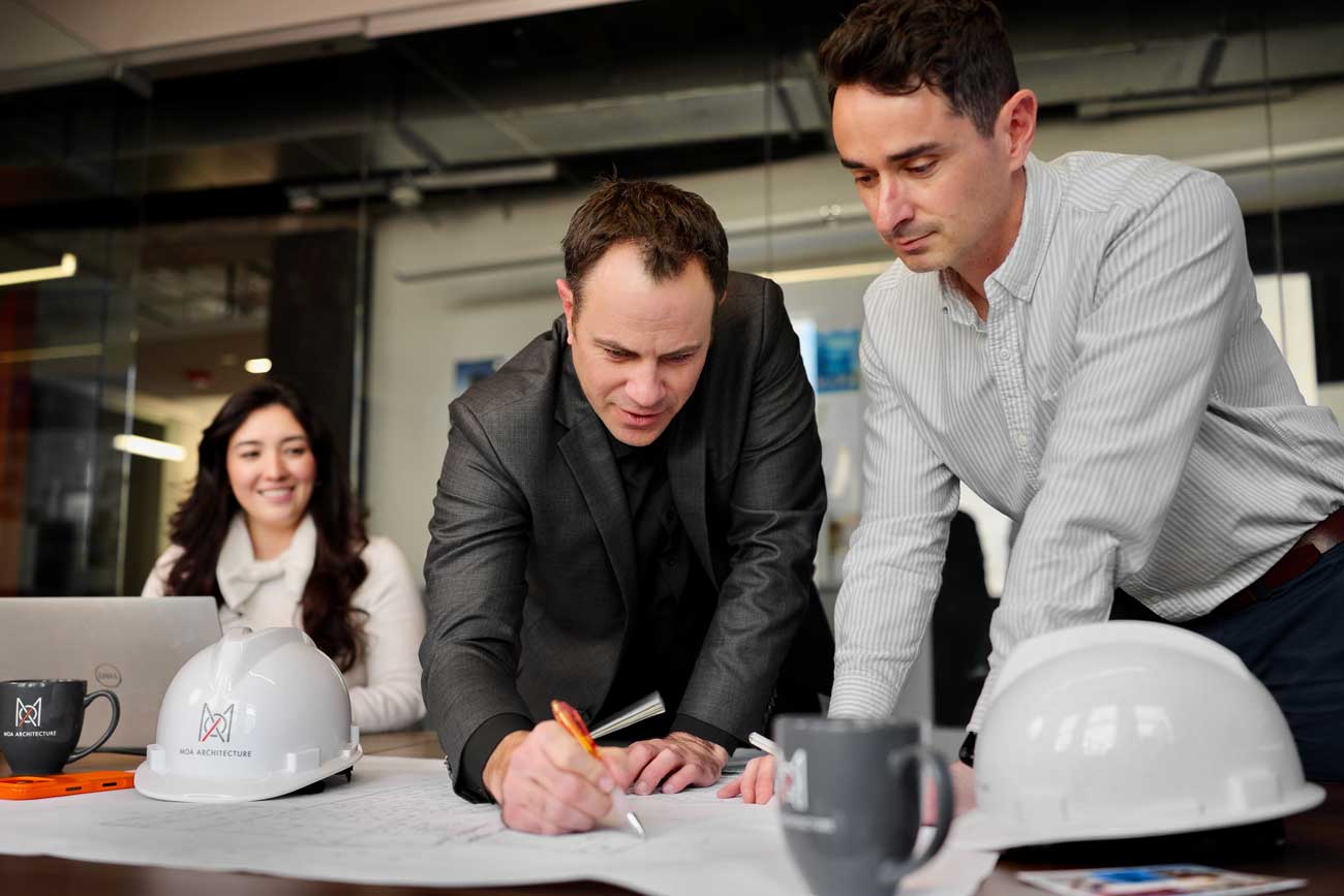 Two MOA Architecture principals reviewing and marking up drawings at a studio table, with MOA-branded hard hats, coffee mugs, and a team member working on a laptop in the background.