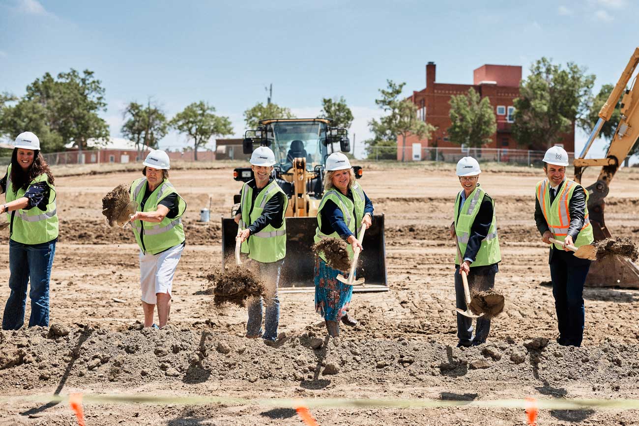 MOA Architecture team members and project stakeholders in hard hats and safety vests tossing shovelfuls of dirt at a groundbreaking ceremony, with construction equipment visible behind them.