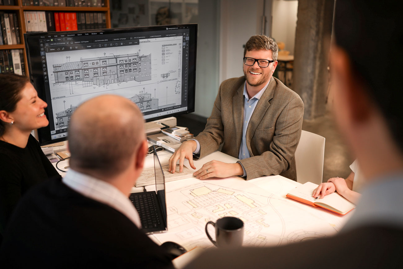 MOA Architecture team members in a design review meeting, smiling and engaged around a table with blueprints and a large monitor displaying architectural construction documents.