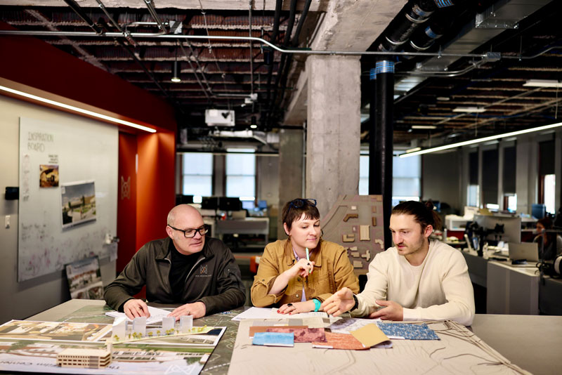 Three MOA Architecture team members reviewing a physical building model, material samples, and drawings at a studio table with an inspiration board visible in the background.