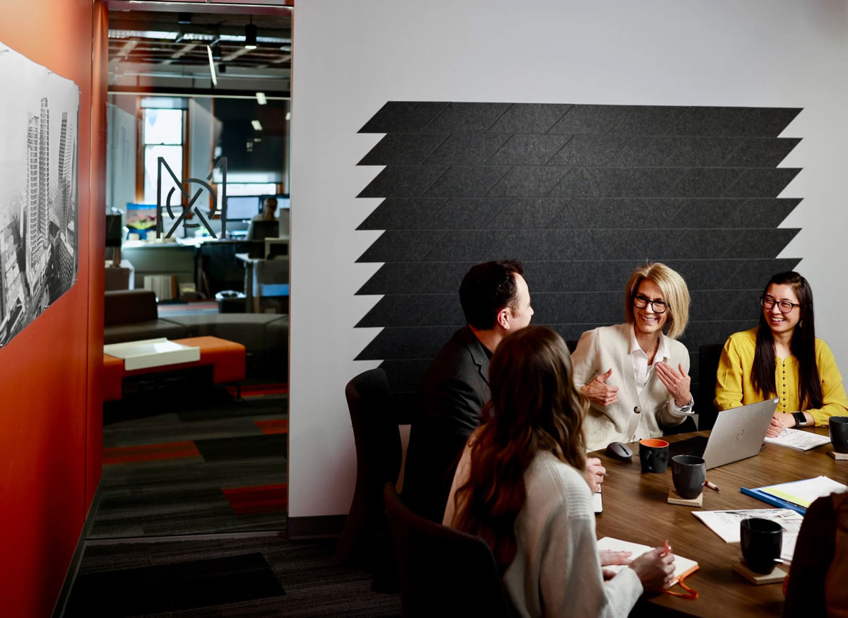 Four MOA Architecture team members in a meeting room with a decorative acoustic wall panel, with the open office lounge visible through the doorway behind them.