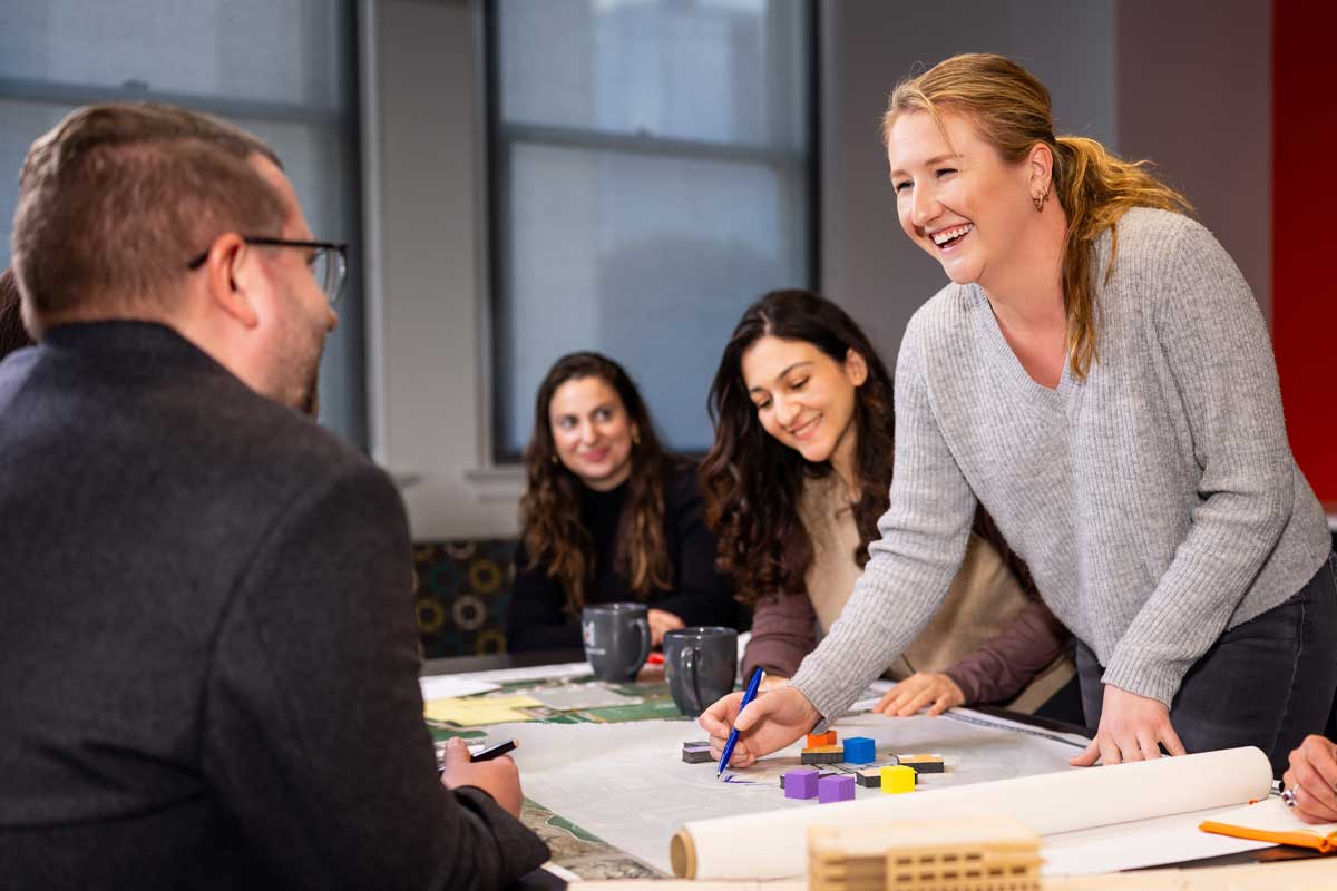 MOA Architecture team members laughing and collaborating around a table with site plans, colorful block models, and rolled blueprints during a design work session.