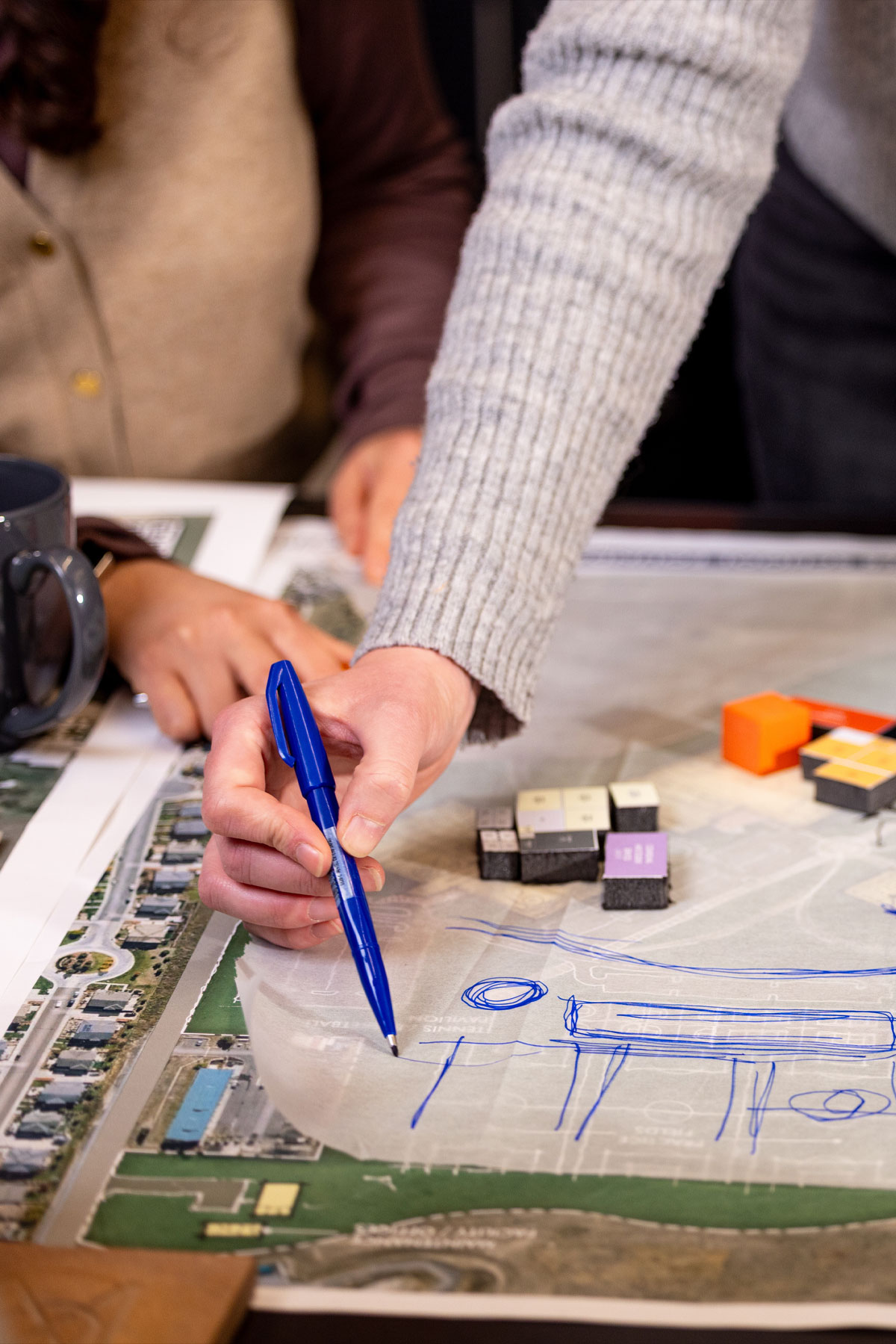 An MOA architect sketching design concepts on tracing paper overlaid on an aerial site map, with small block models nearby representing early planning options.
