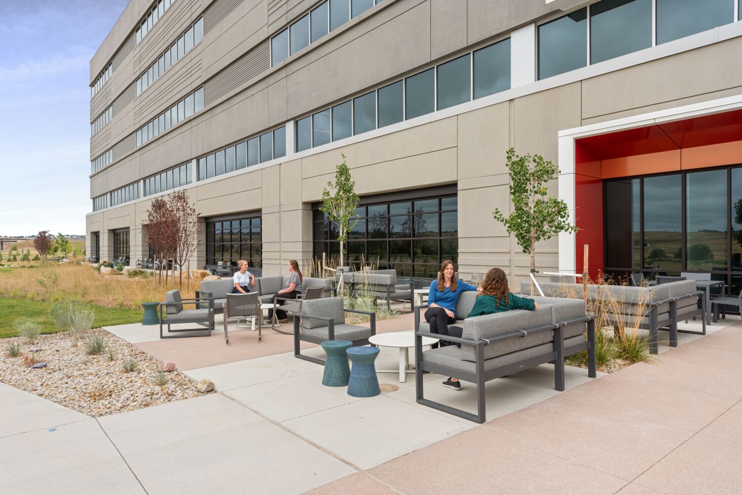 Outdoor lounge area at the Medtronic campus featuring patio seating, small tables, planters with young trees, and the adjacent building façade with large windows.