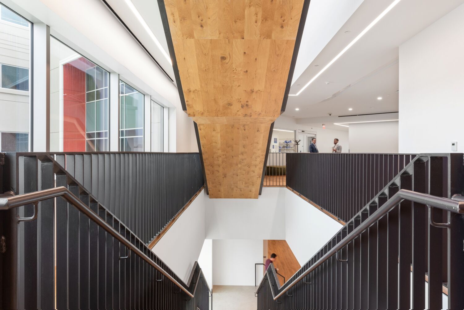 Interior view of a Medtronic workplace showing a central staircase with black metal railings, a wood‑clad underside, white walls, and floor‑to‑ceiling windows along the corridor.