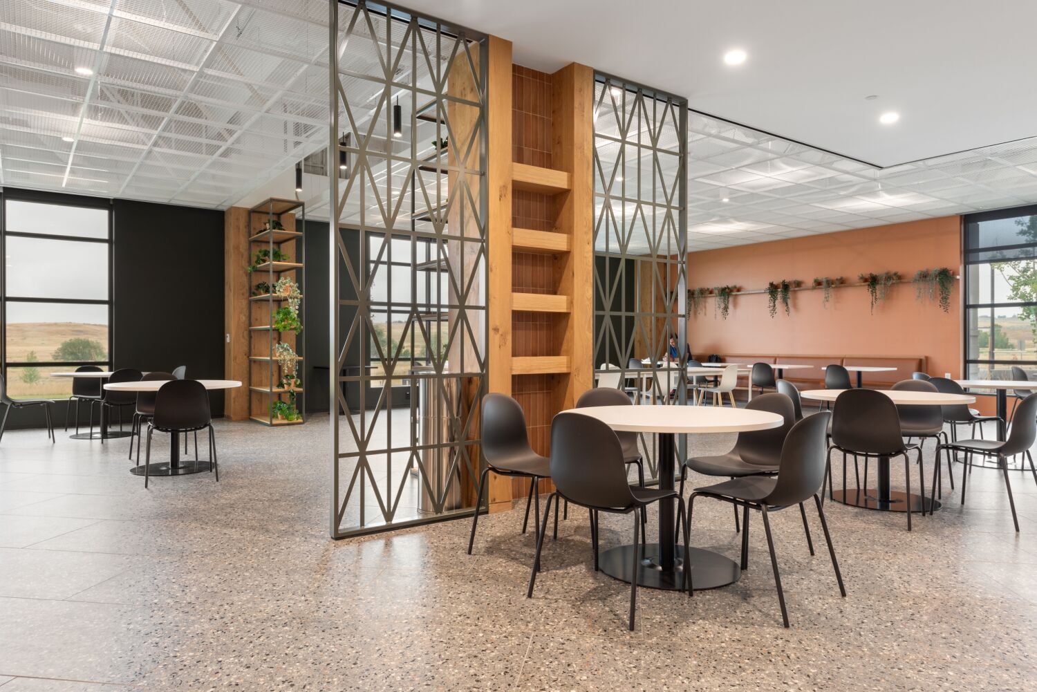Dining space in the Medtronic Marketplace featuring round tables with black chairs, terrazzo flooring, decorative metal screens with wood shelving, a warm orange accent wall with mounted planters, and large windows.