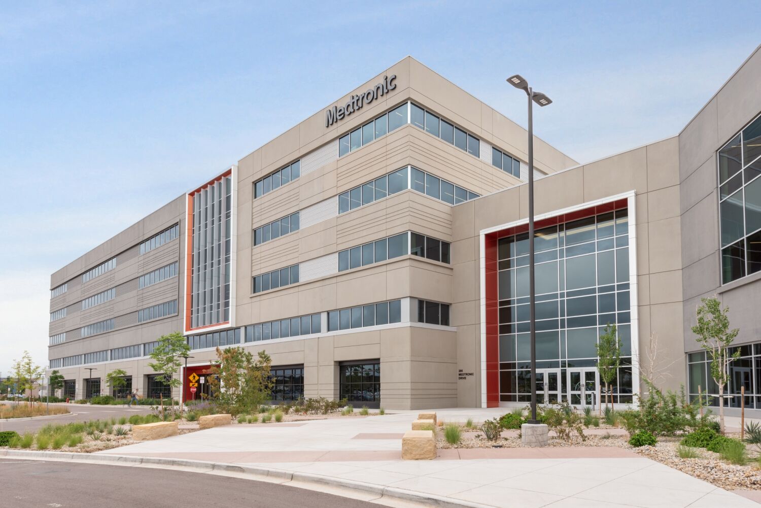 Exterior view of a Medtronic campus building featuring a multi‑story façade with large windows, red accent framing at the main entrance, concrete walkways, and landscaped planting beds.