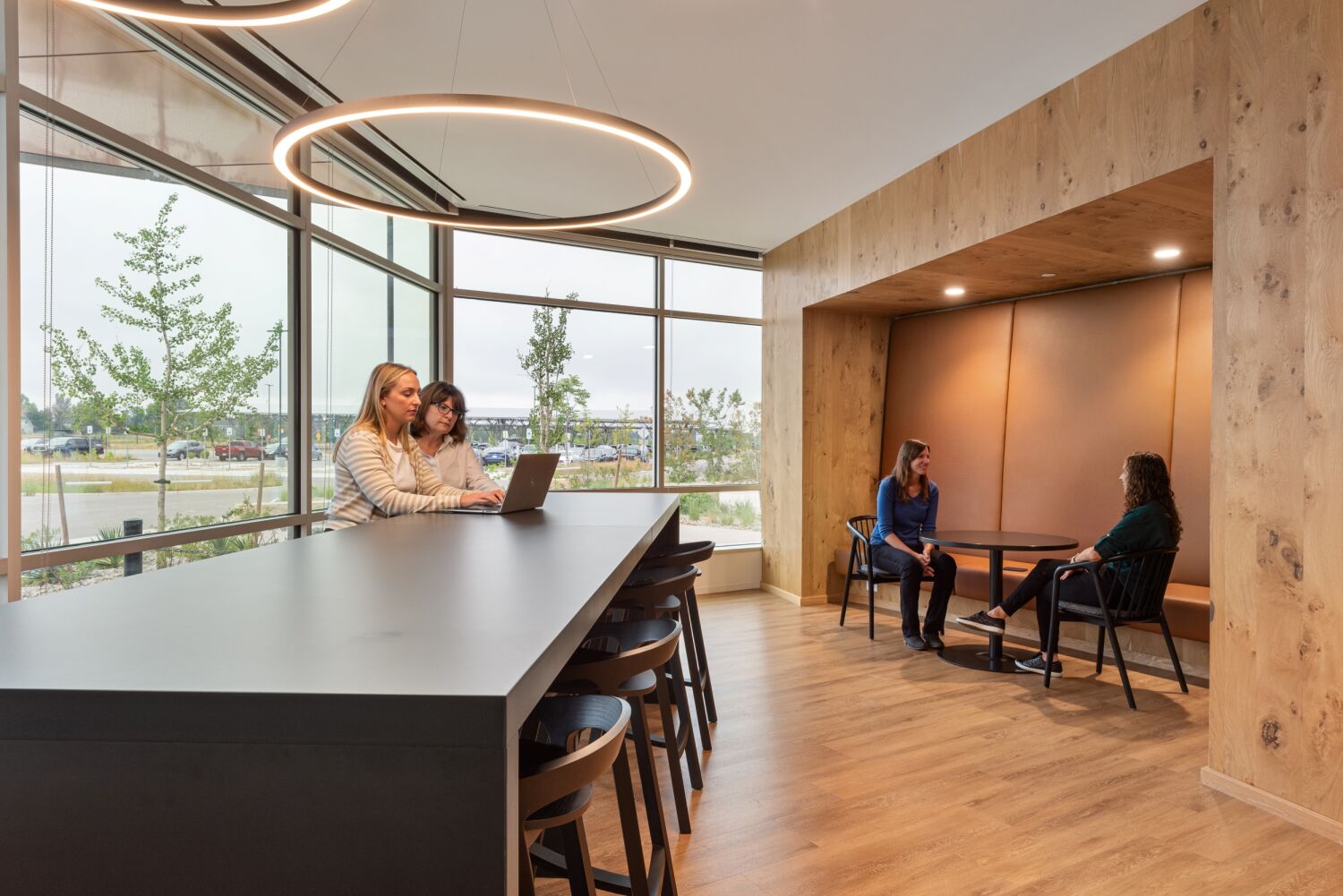 Modern workplace collaboration space at Medtronic featuring a long communal table with ring-shaped pendant lighting, large windows, wood flooring, and a built-in seating nook.