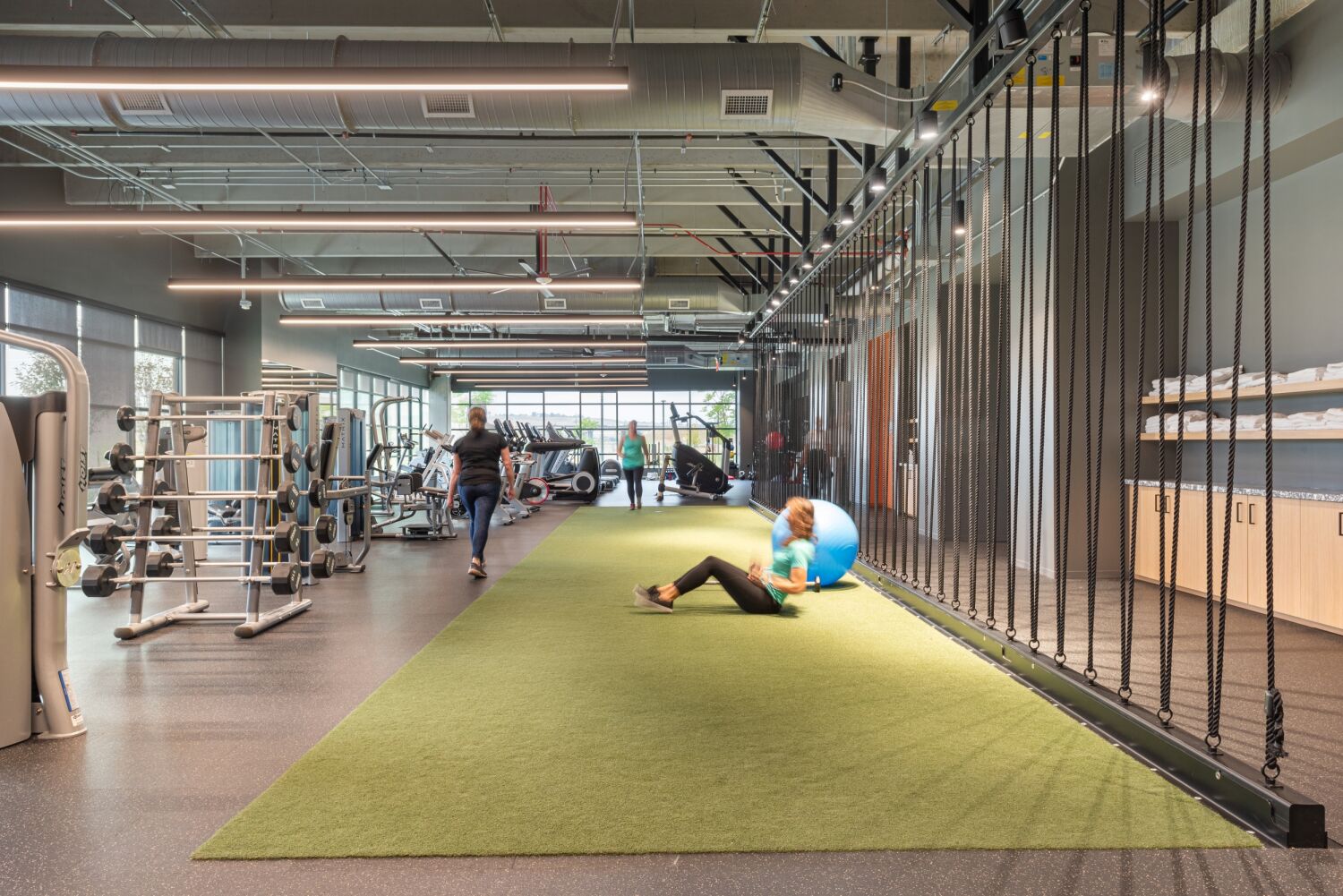 Training turf zone in the Medtronic fitness center featuring strength equipment, rope‑wall stations, large windows, exposed mechanical ceilings, and rubber athletic flooring.