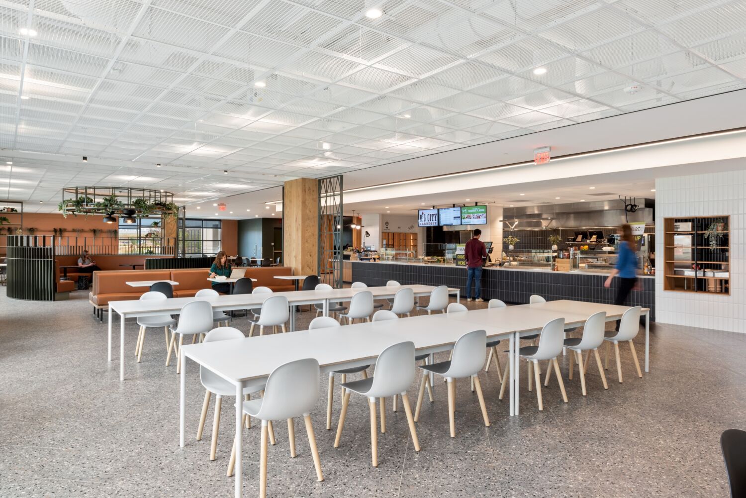 Dining area inside the Medtronic Marketplace featuring long communal tables, booth seating, terrazzo flooring, a greenery chandelier feature, and an open kitchen service counter with menu screens.