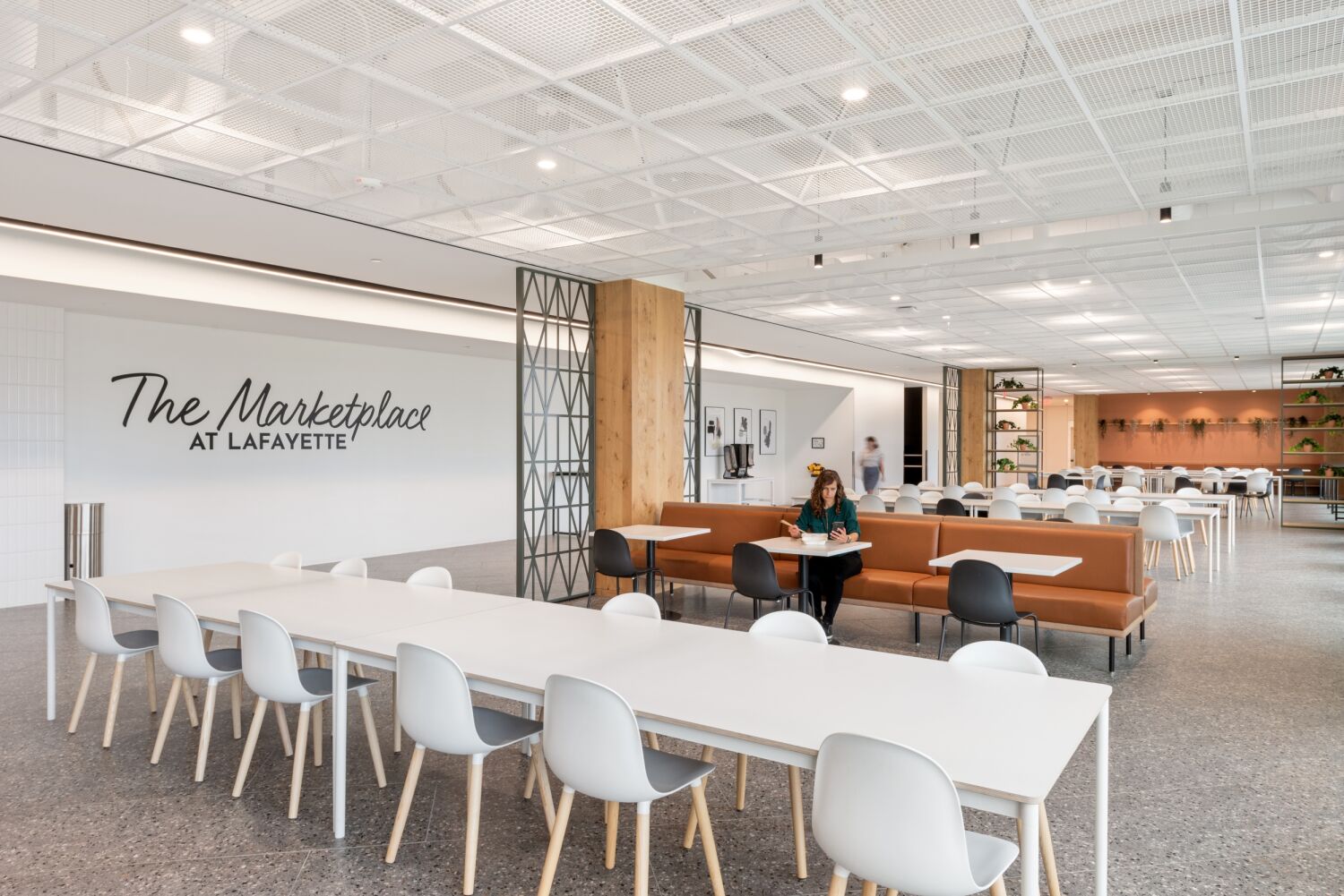 Interior dining hall labeled ‘The Marketplace at Lafayette’ featuring long communal tables, booth seating, wood accent walls, greenery shelving, and a white open‑grid ceiling system.