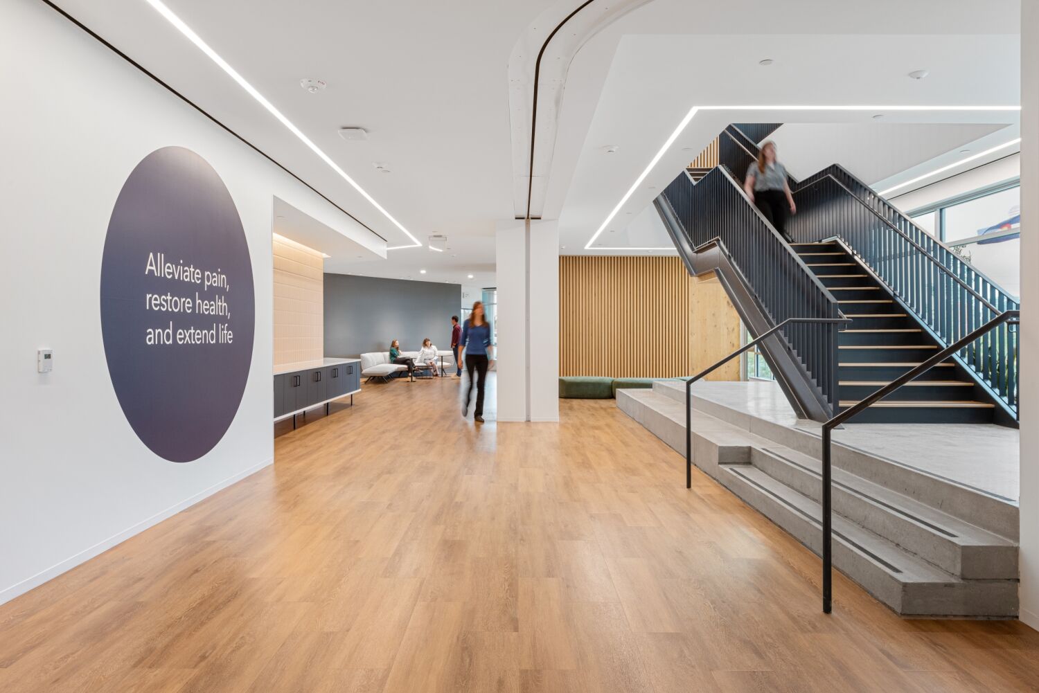 Open workplace lobby at Medtronic featuring wood flooring, a black metal staircase, built‑in seating along a slatted wood wall, and a large circular wall graphic reading ‘Alleviate pain, restore health, and extend life.’