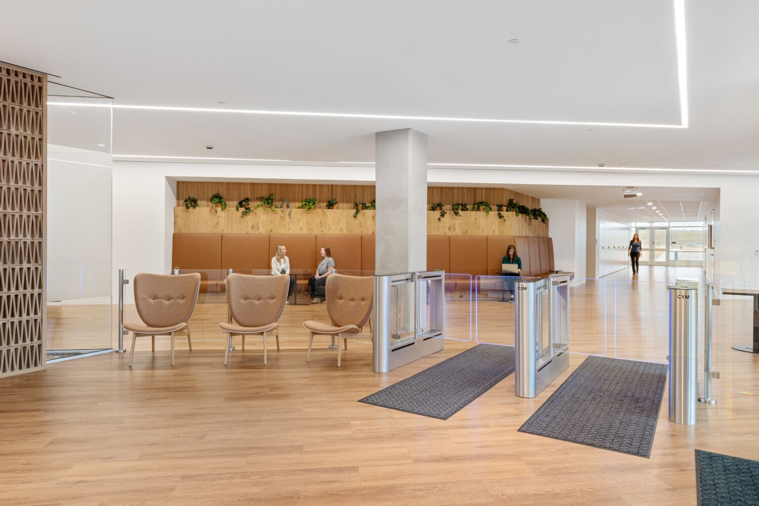 Lobby area in a Medtronic office featuring turnstile security gates, wood flooring, upholstered bench seating with greenery above, and lounge chairs near a central column.