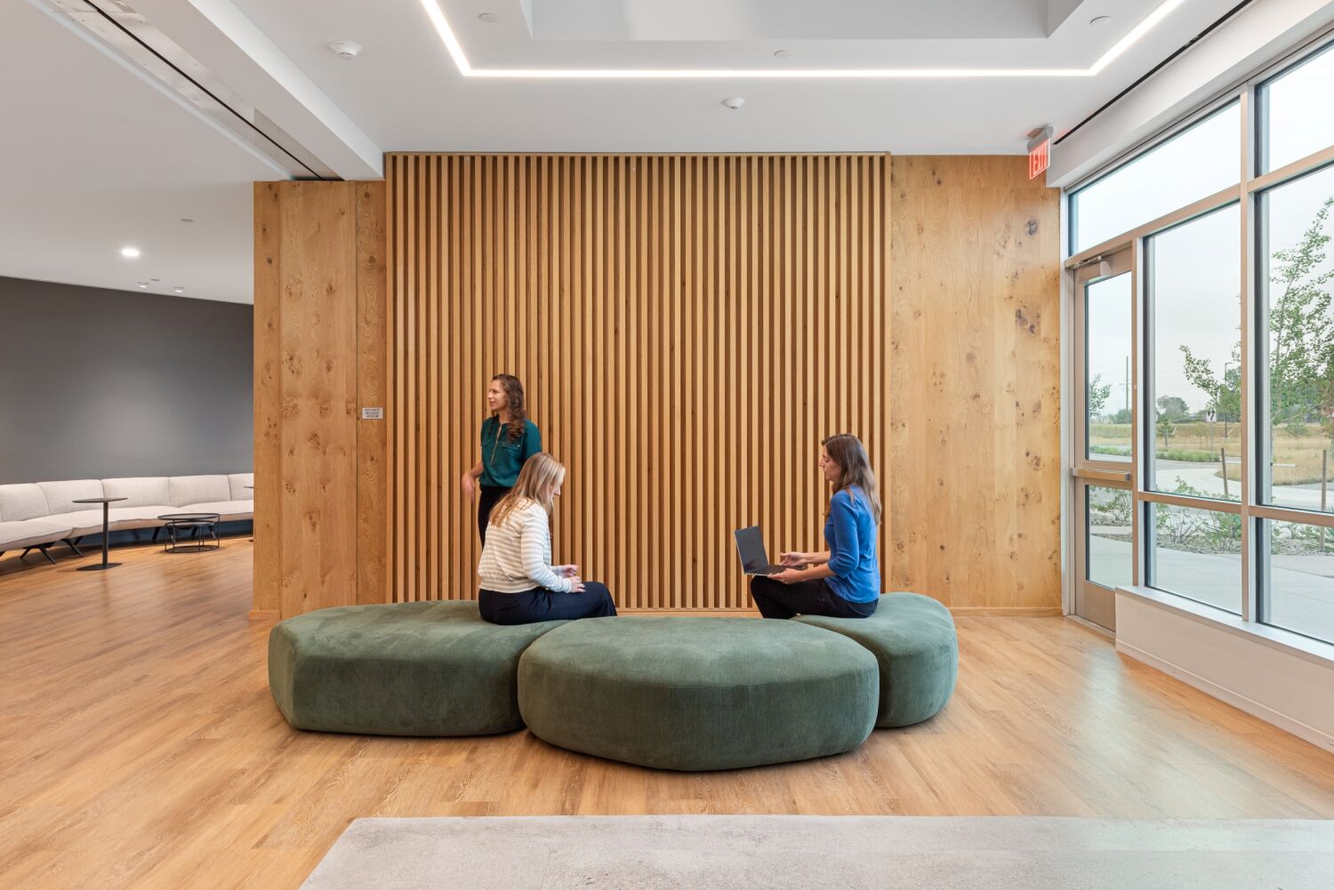 Collaboration lounge in a Medtronic workplace featuring green upholstered seating, a vertical wood‑slat accent wall, wood flooring, and large windows bringing in natural light.