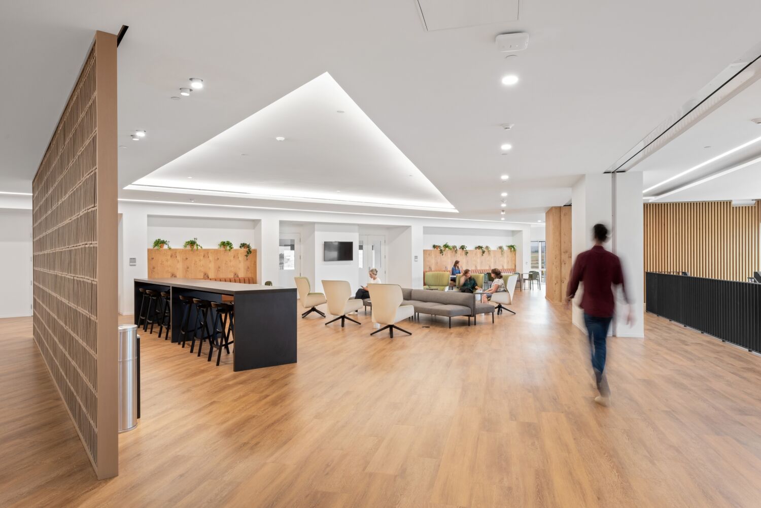 Interior lounge area in a Medtronic office featuring wood flooring, a long high‑top table with stools, lounge seating clusters, a slatted wood accent wall, and recessed ceiling lighting.