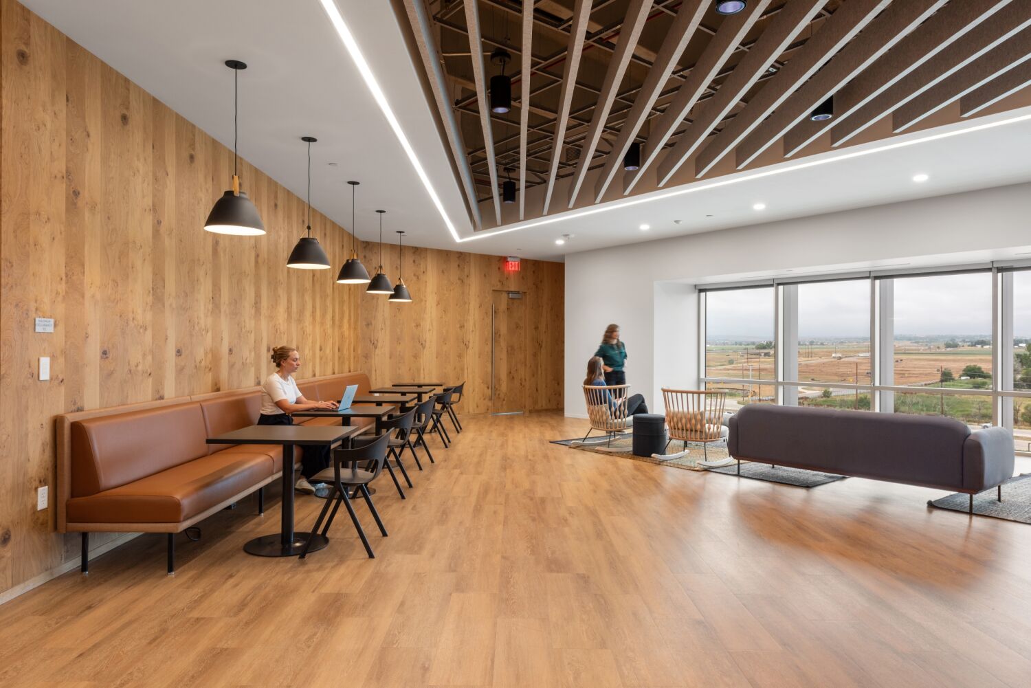 Café‑style lounge area in a Medtronic office featuring wood wall paneling, pendant lighting above booth seating, a slatted ceiling detail, large windows, and wood flooring.