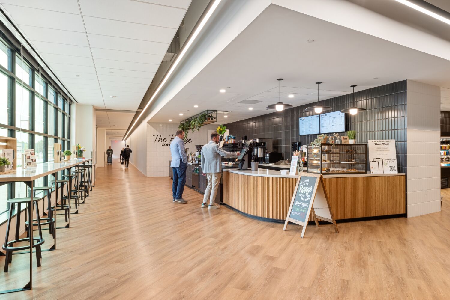 Café service area at the Medtronic campus featuring a curved wood counter, digital menu screens, pendant lighting, black tile wall, and a row of tall windows with bar-height seating.