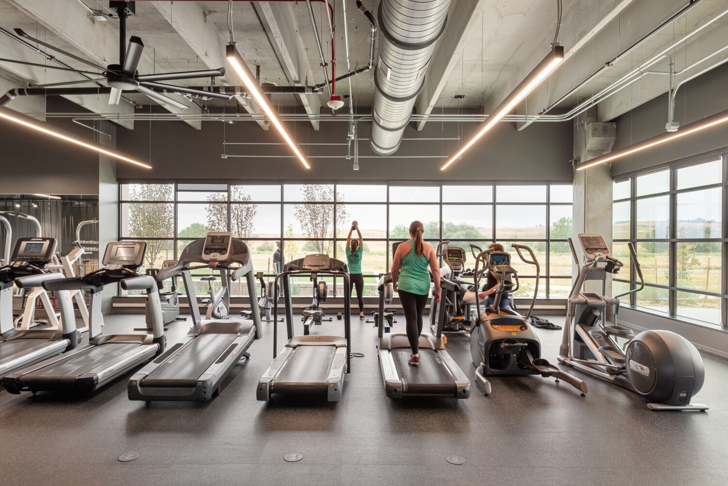 Cardio area in the Medtronic fitness center featuring treadmills, elliptical machines, large windows overlooking the landscape, exposed ductwork, and linear ceiling lights.