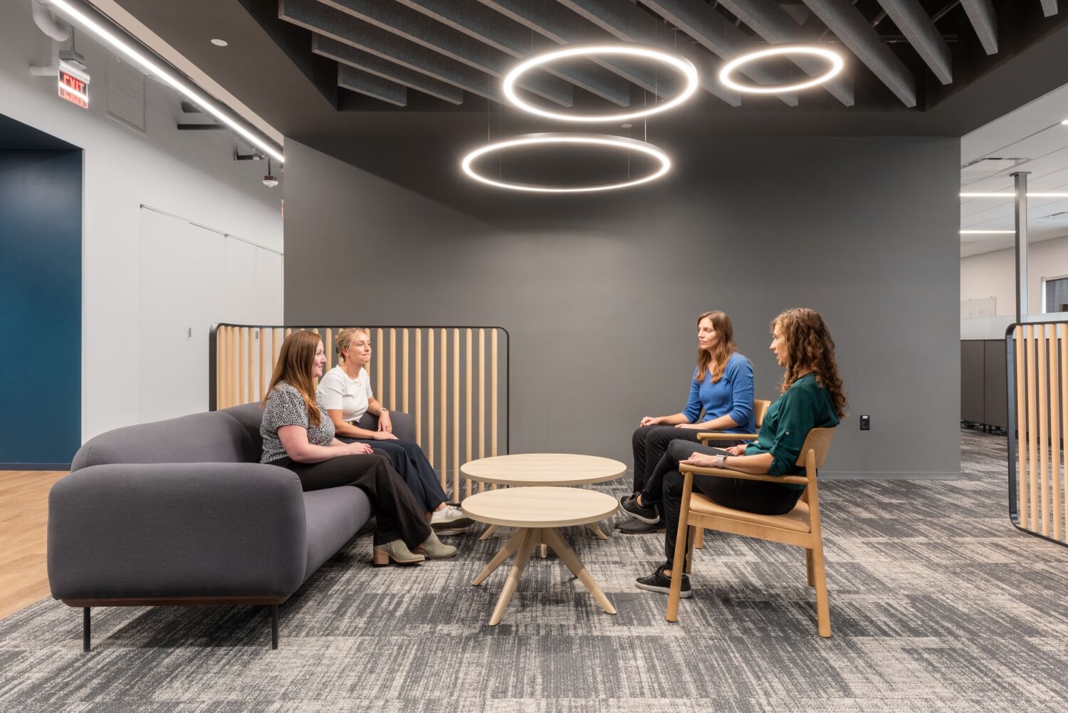 Collaboration lounge in a Medtronic workplace featuring a gray sofa, round wooden tables, wood‑slat partition panels, circular pendant lights, acoustic ceiling baffles, and patterned carpet flooring.