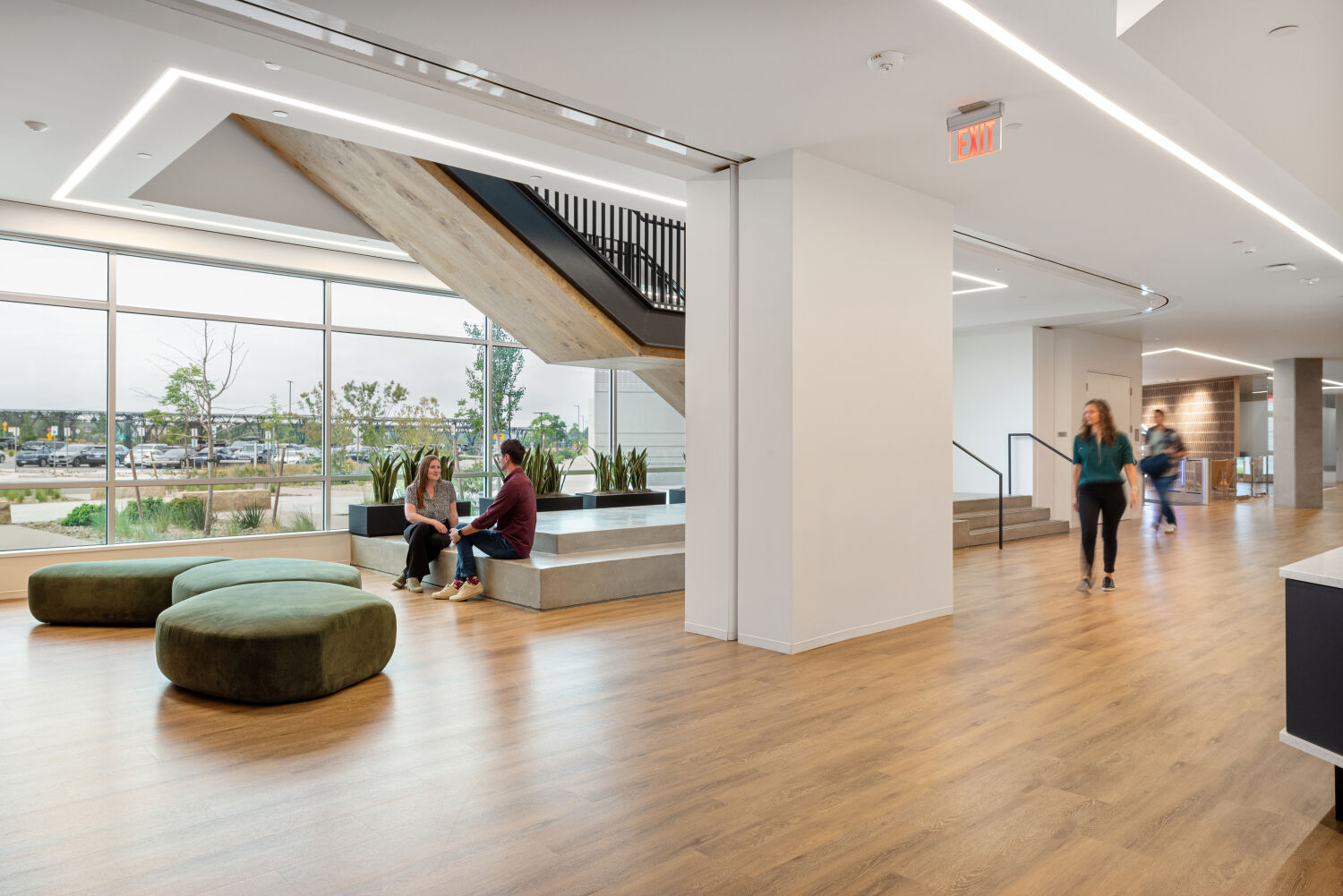 Open atrium space in a Medtronic workplace featuring concrete tiered seating beneath a large staircase, wood flooring, floor-to-ceiling windows, and green upholstered lounge seating.