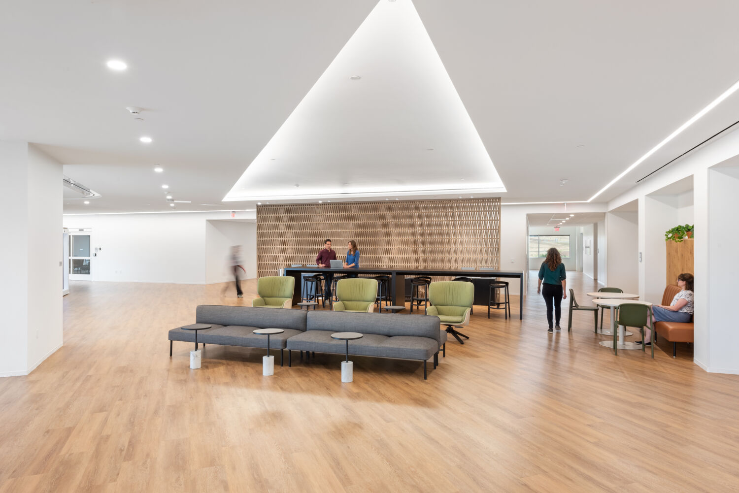 Open interior shared lounge at a Medtronic workplace featuring wood flooring, a patterned feature wall behind high‑top tables, lounge seating clusters, and recessed ceiling lighting.