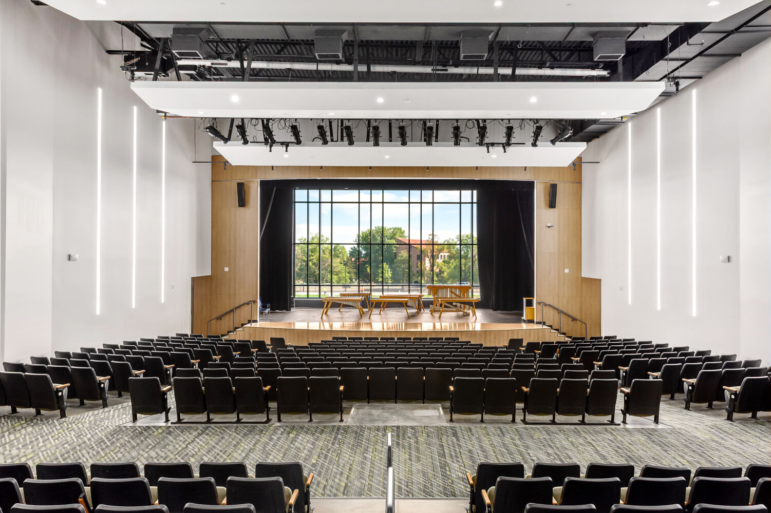 Auditorium with tiered seating, patterned carpeted aisles, a proscenium stage framed by wood‑accent panels, and full‑height glazing behind the stage revealing outdoor views.