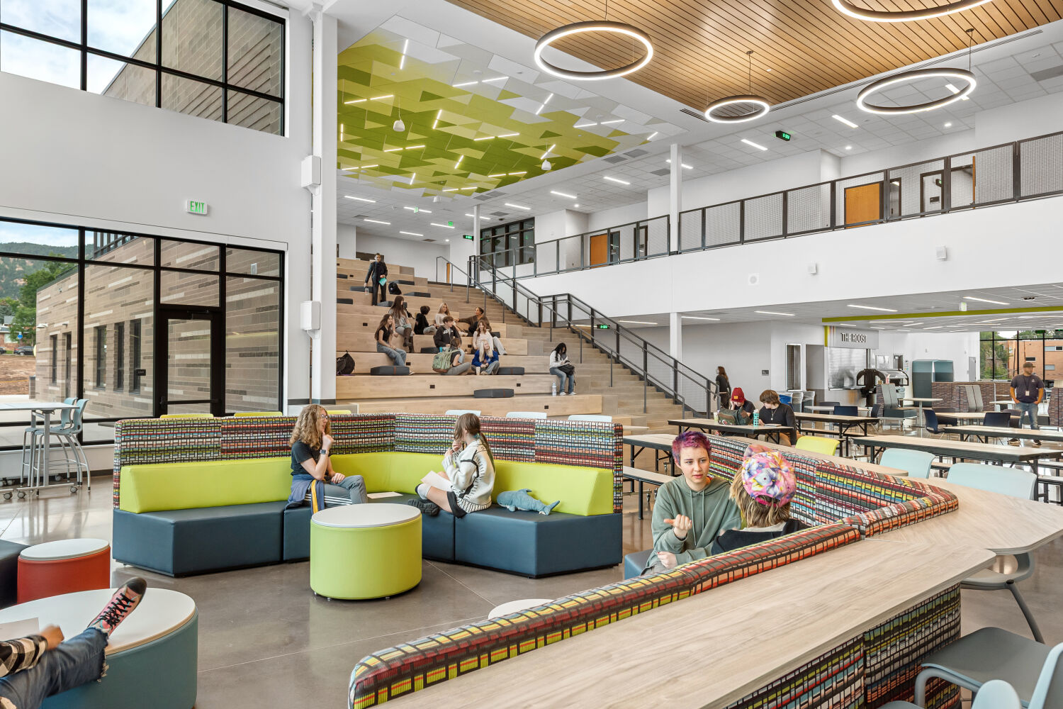 Commons with curved lounge seating in patterned upholstery, polished concrete flooring, cafeteria tables, a tiered learning stair with wood seating, large windows, and a ceiling with geometric green acoustic panels and circular pendant lights.