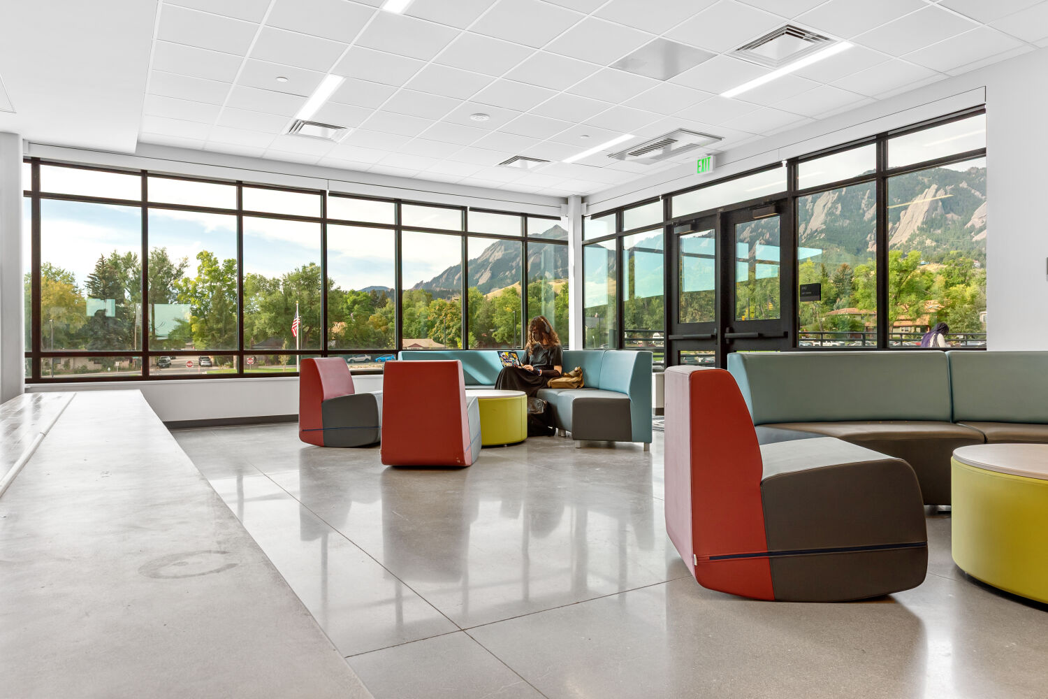 Commons lounge area with colorful modular seating, polished concrete flooring, floor‑to‑ceiling windows with mountain views, and a ceiling grid with recessed lighting.