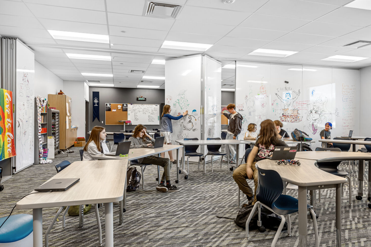 Classroom with movable tables and chairs, floor‑to‑ceiling writable wall panels covered with drawings and notes, patterned carpet tile flooring, storage cabinets, and a suspended ceiling with recessed lighting.