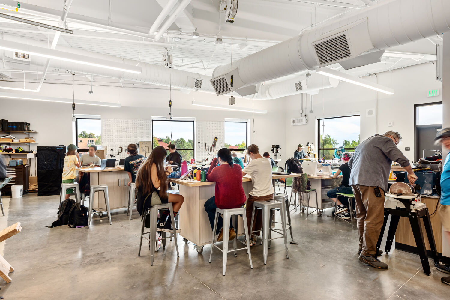 Fabrication lab with workbenches, metal stools, pegboard tool walls, large windows, exposed ductwork, overhead task lighting, storage shelves, and various equipment and materials arranged throughout the space.