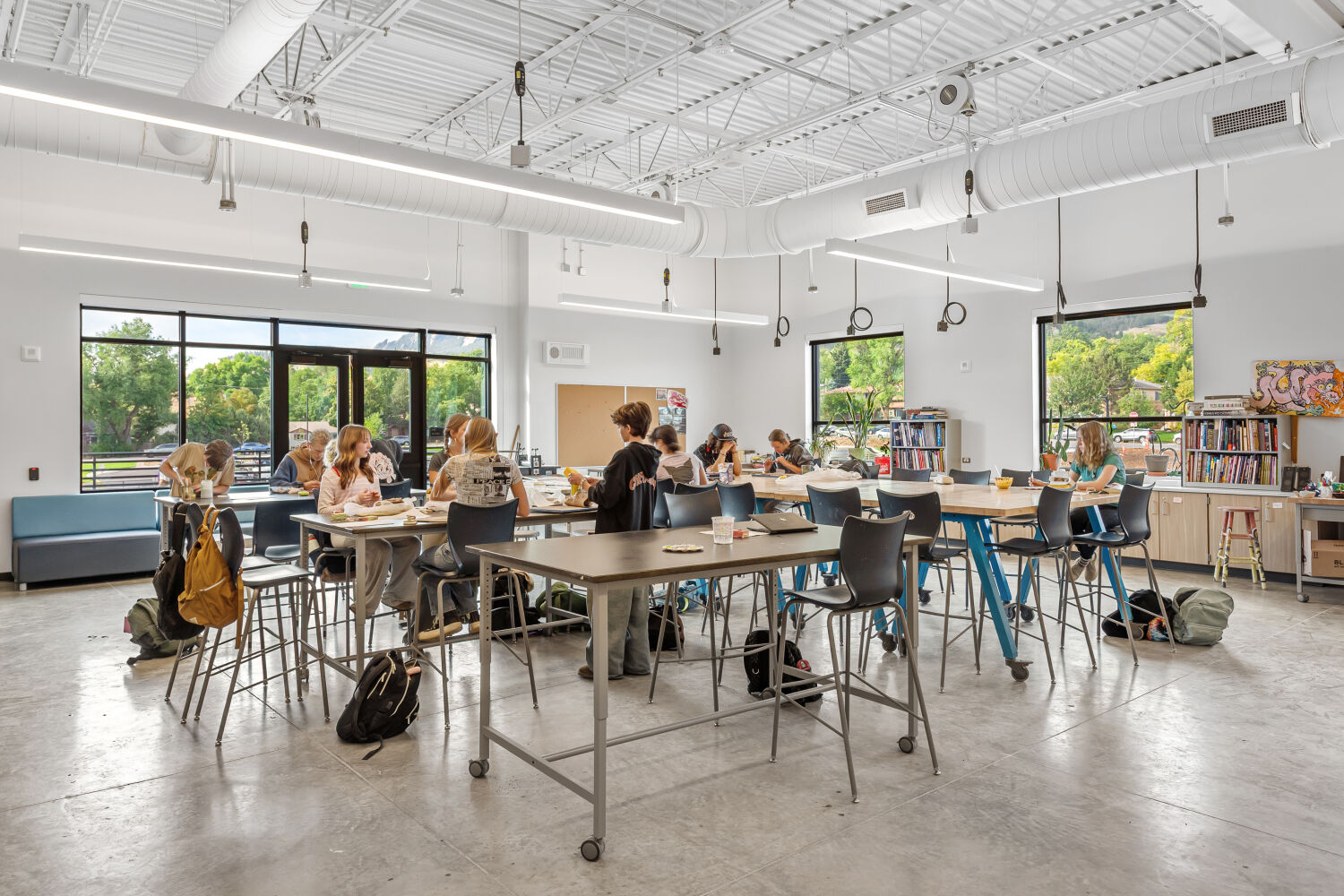 Art classroom with high worktables and stools, large windows providing natural light, open shelving with art supplies, pegboard storage walls, and concrete flooring.