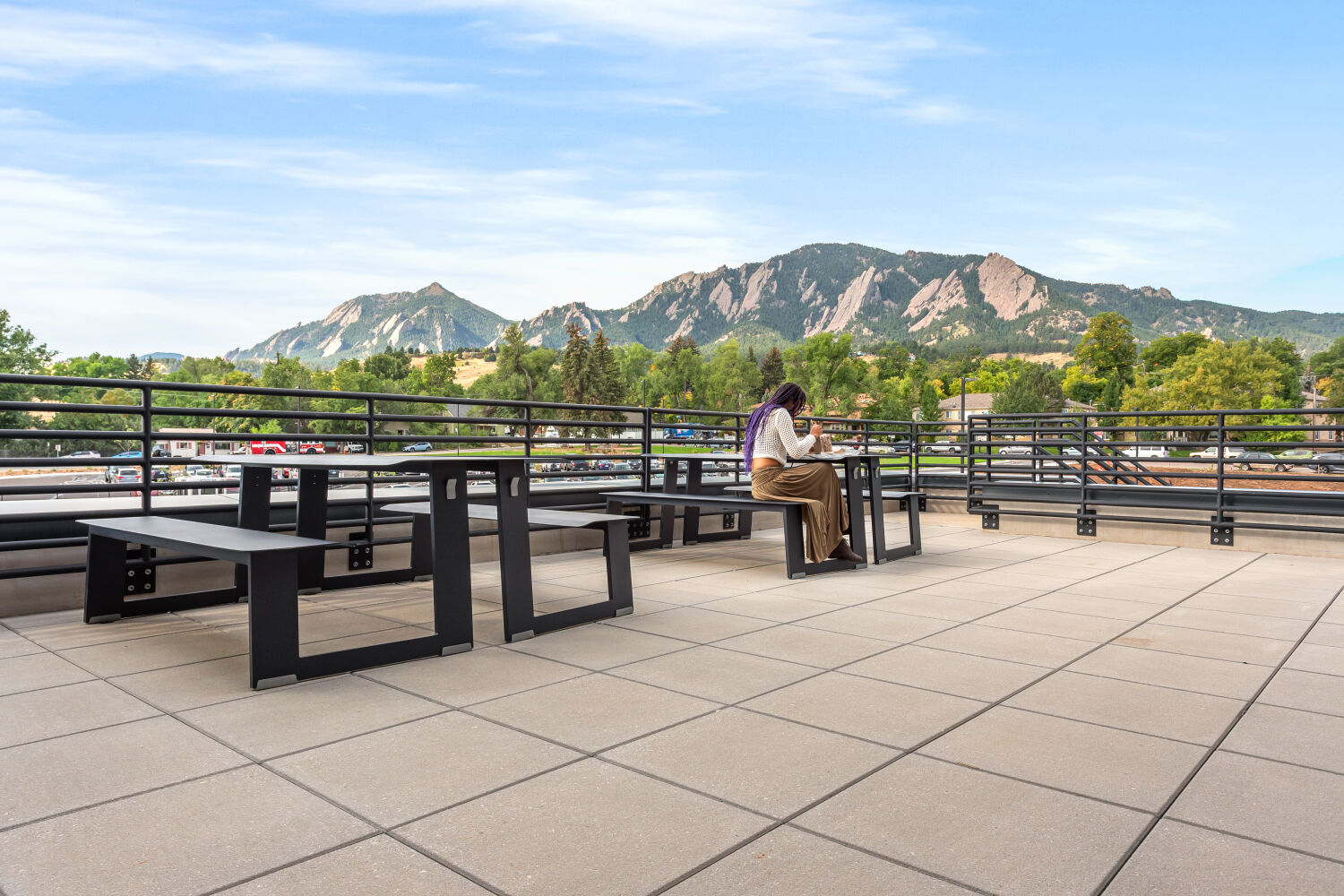 Outdoor art patio with metal tables and benches, tiled flooring, perimeter railing, and expansive mountain views under a clear sky.