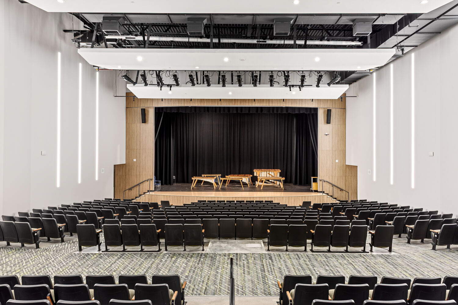 Auditorium with tiered seating, patterned carpeted aisles, a proscenium stage with black curtains, wood‑accent wall panels, overhead stage lighting, and a ceiling with suspended acoustic panels and linear lighting.