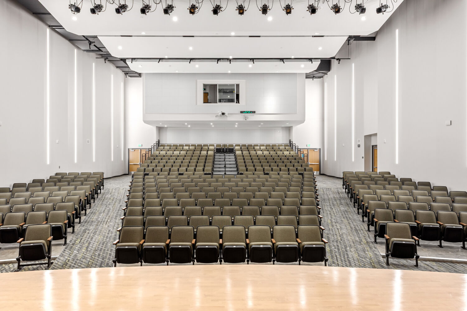 Auditorium viewed from the stage, featuring tiered seating with upholstered chairs, patterned carpeted aisles, side walls with vertical linear lighting, and a ceiling with suspended acoustic panels and stage lighting fixtures.