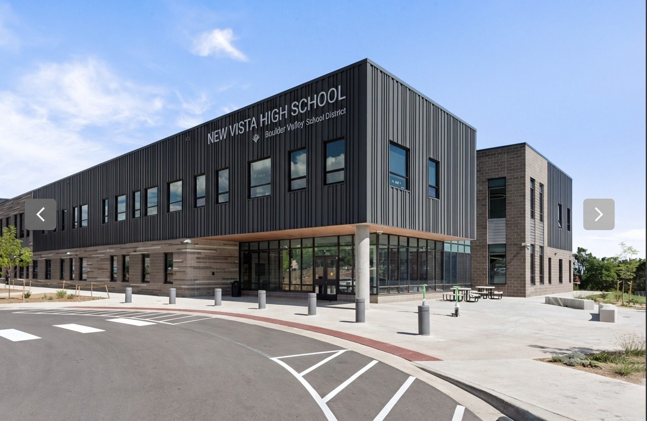 Exterior view of a modern high school building with vertical metal panel cladding, large windows, a cantilevered second‑floor massing, ground‑level glass entry, concrete columns, and adjacent outdoor seating areas.