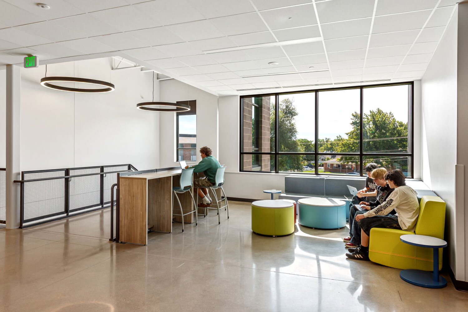 Collaborative seating area with a high‑top counter, bar‑height stools, circular lounge seats, large windows with exterior views, polished concrete flooring, and circular pendant lights.