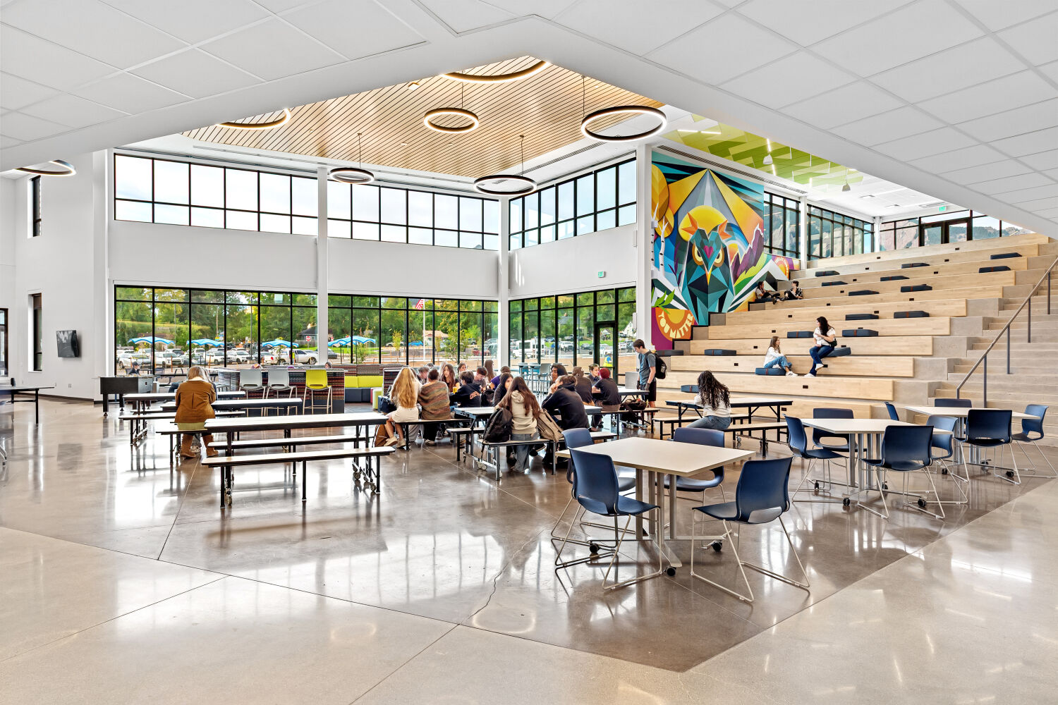 Open two‑story commons with polished concrete flooring, floor‑to‑ceiling windows, wood‑slat ceiling accents with circular pendant lights, cafeteria tables, a tiered learning stair, and a large colorful geometric mural on the adjacent wall.