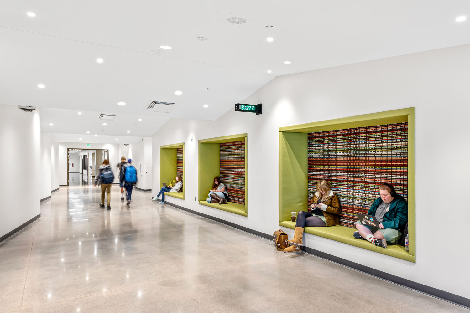 School corridor with large recessed seating alcoves featuring patterned back panels and green cushions, polished concrete flooring, and white walls with integrated lighting.