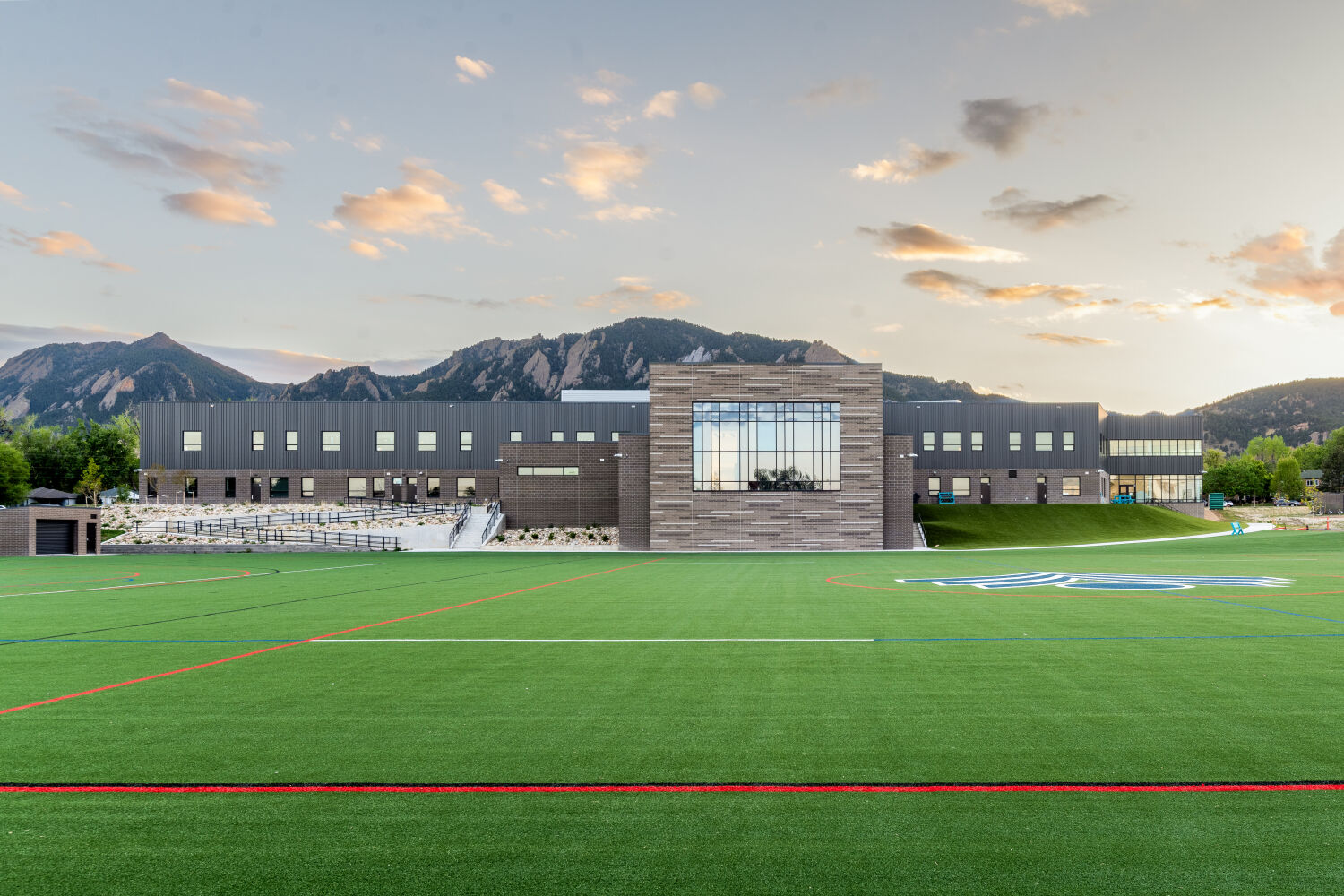 New Vista High School building viewed from an athletic field, featuring a central glazed volume, metal and brick facade, stepped site walls, and adjacent turf field with court markings.