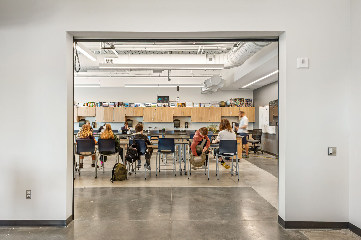 Wide interior opening looking into a classroom with high worktables and stools, wood cabinetry along the back wall, pegboard tool storage, exposed ductwork, overhead linear lighting, and polished concrete flooring.