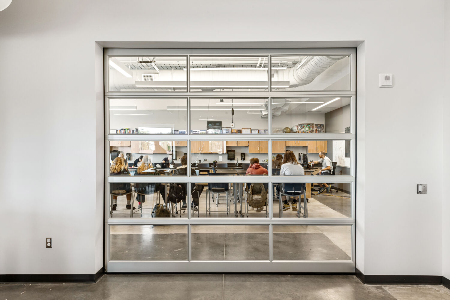 View through a large interior glass garage‑style door into a classroom with high tables, stools, wood cabinetry, pegboard tool walls, exposed ductwork, overhead lighting, and concrete flooring.
