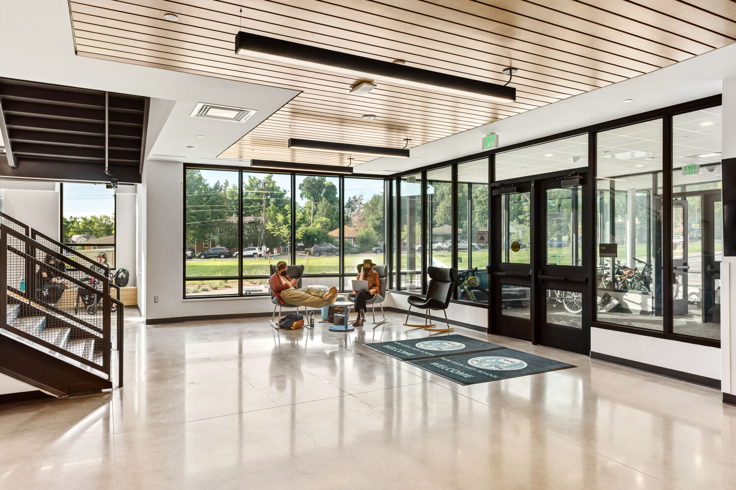 Front entrance lobby with large floor‑to‑ceiling windows, glass entry doors, wood‑slat ceiling accents, polished concrete flooring, modern lounge seating, and an open stair with metal railing.