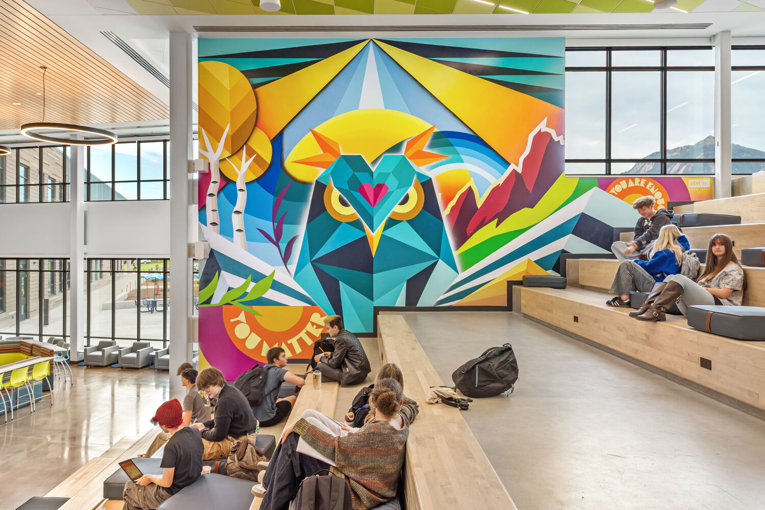 Learning stair with tiered wood seating, adjacent concrete steps with metal handrails, a large geometric owl mural in bright colors, polished concrete flooring, expansive windows, and a ceiling with green acoustic panels.