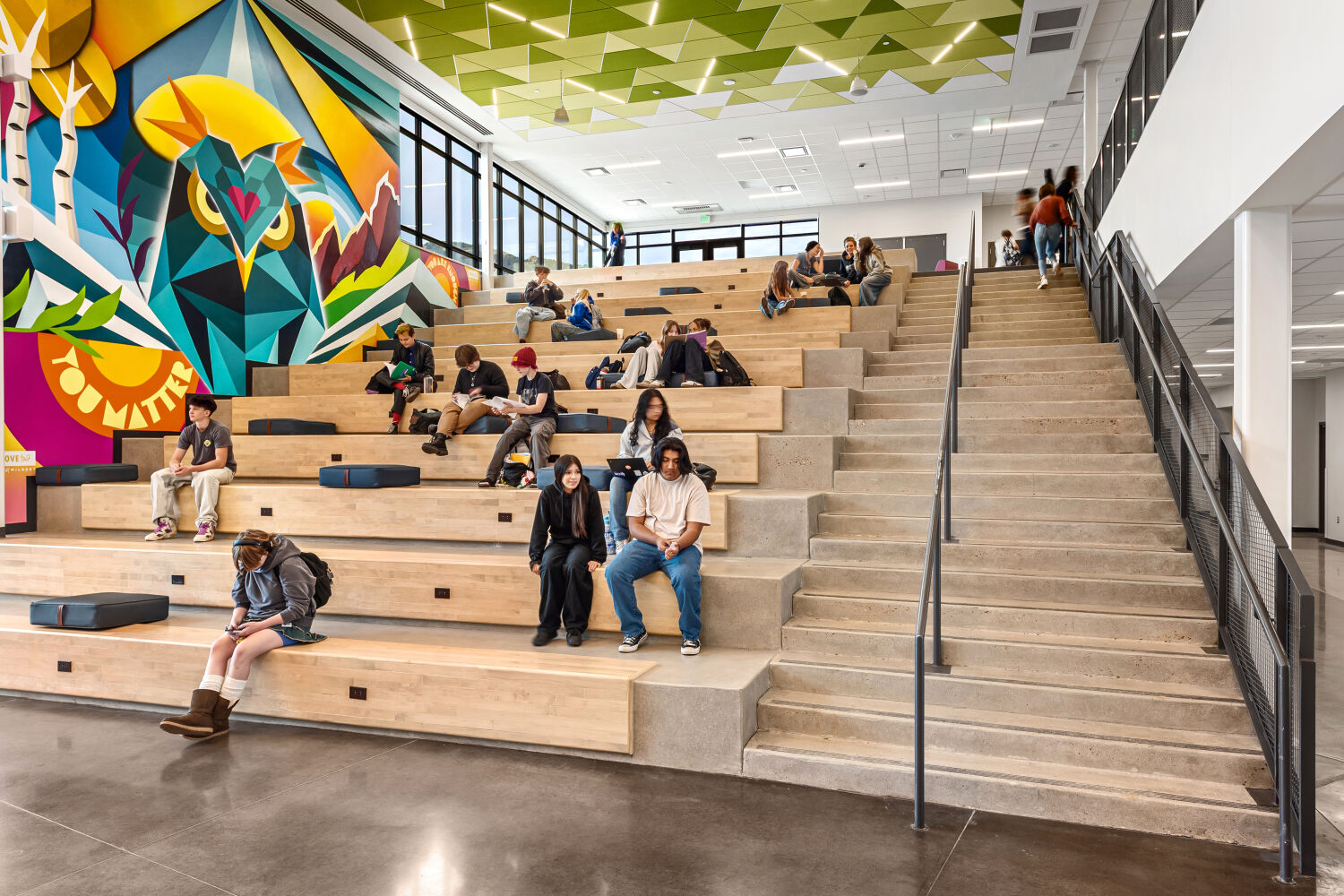 Learning stair with tiered wood seating, adjacent concrete steps with metal handrails, large geometric mural in bright colors, polished concrete flooring, and expansive windows providing natural light.